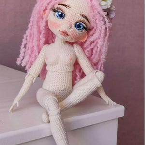 May include: A crocheted doll with pink yarn hair and a flower crown sits on a white surface. The doll has blue eyes and a white crocheted body.