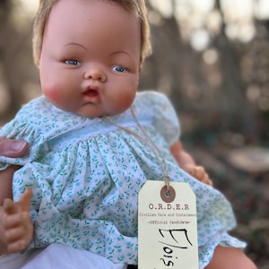 May include: A vintage baby doll with blue eyes and a light blue floral dress. The doll is holding a tag that reads "O.R.D.E.R. Civilian Care and Containment -Official Candidate- Eloise O.R.D.E.R. Official Doll"