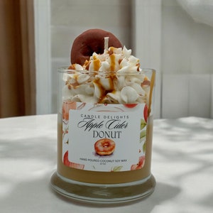 May include: A clear glass candle with a caramel-colored wax base, topped with white whipped wax, a brown donut, and caramel drizzle. The label reads "Candle Delights Apple Cider DONUT" with a donut illustration. The candle is 12 oz.