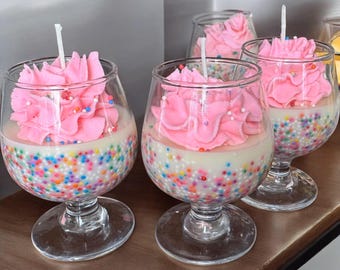 May include: Four glass candle holders filled with white wax and topped with pink frosting-like candle decorations. The candles are sprinkled with colorful sprinkles.
