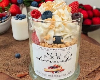 Könnte beinhalten: Eine Kerze im Klarglasgefäß, verziert mit weißer Wachs-Schlagsahne, blauen Wachs-Heidelbeeren, roten Wachs-Himbeeren und beigen Wachskrümeln. Das Etikett trägt die Aufschrift KC DESIGNS, CO. WILD BERRY cheesecake ALL NATURAL HAND POURED SOY CANDLE.