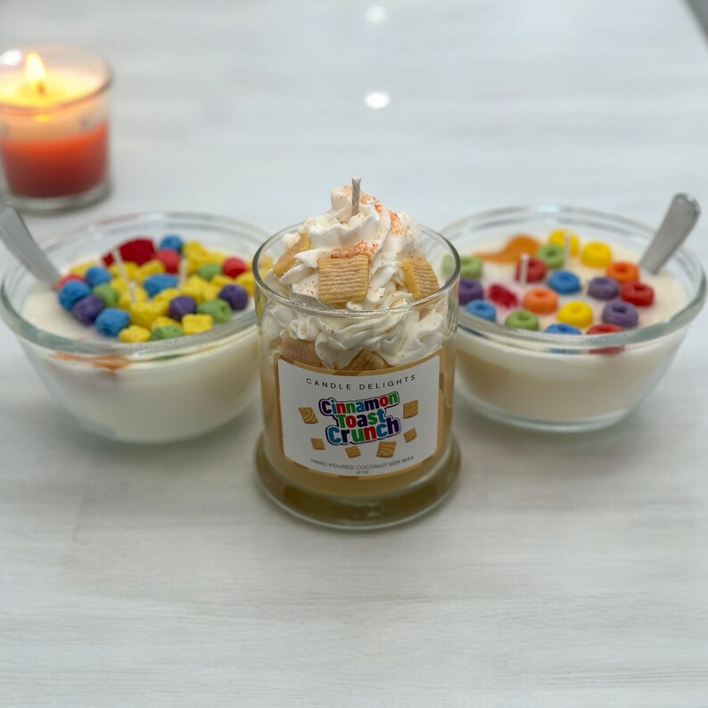 May include: Three glass jars with white wax and colorful cereal pieces. The center jar has a label that reads "Candle Delights Cinnamon Toast Crunch Hand Poured Coconut Soy Wax".