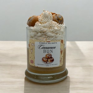 May include: A glass jar candle with a white frosting-like topping and a brown cinnamon bun on top. The label reads "Candle Delights Cinnamon Bun Hand Poured Coconut Soy Wax Candle 12 oz."