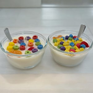 May include: Two glass bowls filled with white wax and colorful cereal-shaped candles. The bowls are on a light wood surface.