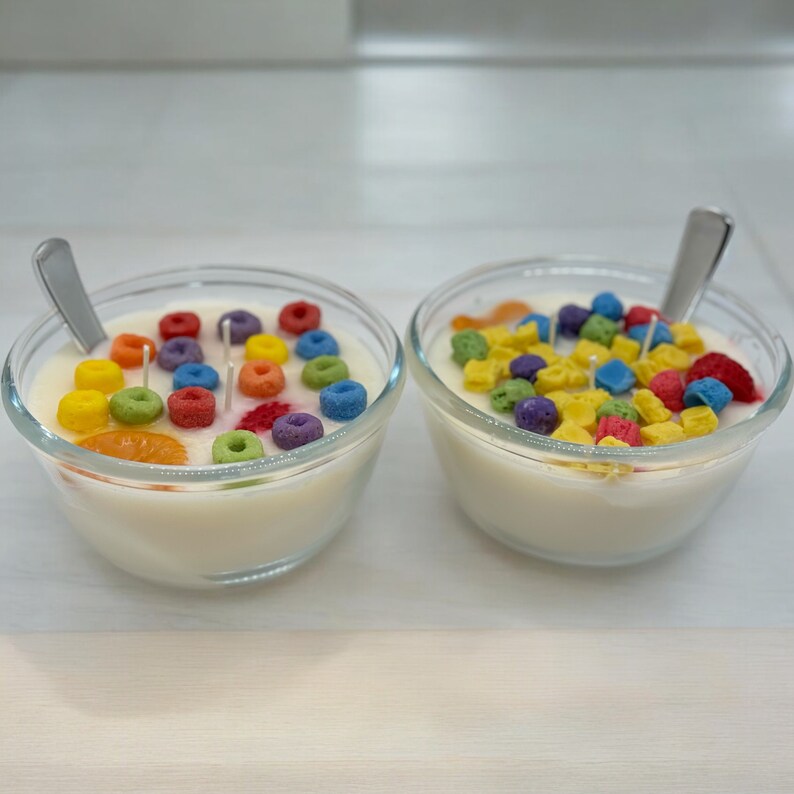 May include: Two glass bowls filled with white wax and colorful cereal-shaped candles. The bowls are on a light wood surface.