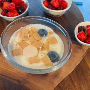 May include: A clear glass jar candle with a white wax base and a variety of toppings. The toppings include small, square, light brown pieces of cereal, two blue blueberry-shaped candles, and a single pink round candle.