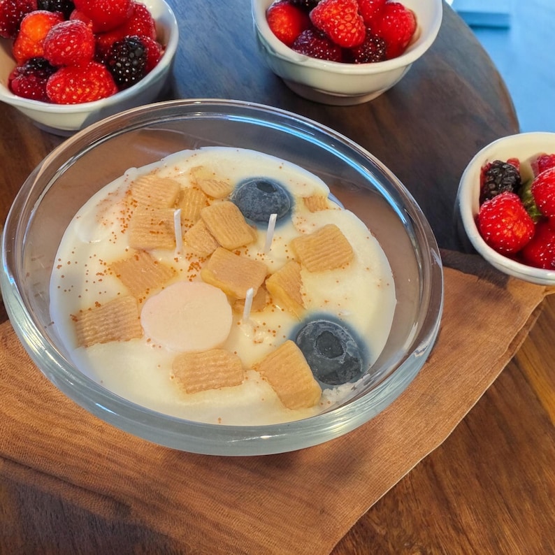 May include: A clear glass jar candle with a white wax base and a variety of toppings. The toppings include small, square, light brown pieces of cereal, two blue blueberry-shaped candles, and a single pink round candle.