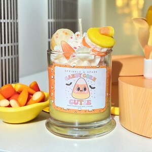 May include: A decorative candle in a glass jar with a candy corn theme. The candle has a yellow base, orange middle, and white top with decorative toppings. The label reads "Candy Corn Cutie" with a candy corn graphic and bats.