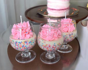 May include: Three pink and white cupcake candles in glass holders with colorful sprinkles. The candles are in the shape of cupcakes with pink frosting and white sprinkles.