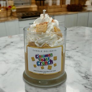 May include: A candle in a glass jar with a white whipped cream topping and cinnamon toast crunch cereal pieces. The label reads "Candle Delights Cinnamon Toast Crunch Hand Poured Coconut Soy Wax 12 oz."