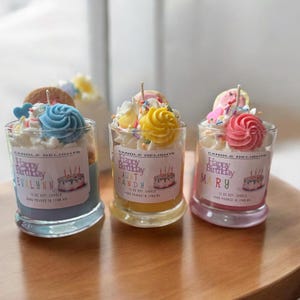 May include: Three small glass jars with colorful birthday candles. Each jar has a different name on the label: "Evalynn", "Aunt Candy", and "Mary". The jars are decorated with frosting, sprinkles, and a small cake.