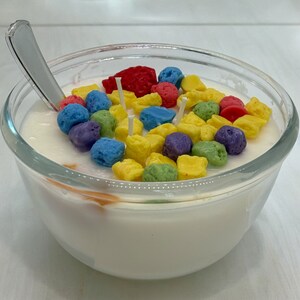 May include: A glass bowl filled with a white substance resembling milk or yogurt, topped with colorful cereal-shaped candles. The candles are in various colors, including yellow, red, blue, green, and purple.