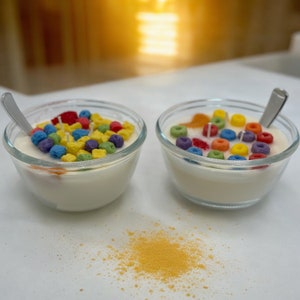 May include: Two glass bowls filled with white wax and colorful cereal-shaped candles. A small pile of yellow wax is scattered on the white surface.