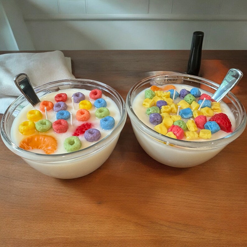 May include: Two clear glass bowls filled with white wax and colorful cereal-shaped candies. The bowls are on a wooden surface.