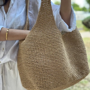 May include: A large, open-weave tote bag in a natural tan colour. The bag has two long straps and a spacious interior. The texture of the bag appears to be woven, and the bag is being held up by a person.