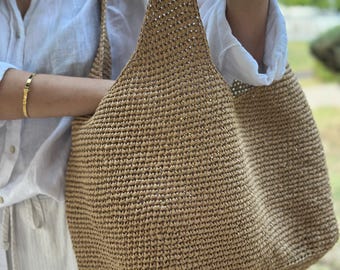 May include: A large, open-weave tote bag in a natural tan colour. The bag has two long straps and a spacious interior. The texture of the bag appears to be woven, and the bag is being held up by a person.