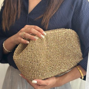 Può includere: Una pochette dorata intrecciata di forma rettangolare. La borsa ha una superficie strutturata ed è tenuta da una persona che indossa una camicia blu navy e pantaloni grigi. La pochette è un accessorio elegante.