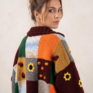 Puede incluir: Un cárdigan de punto grueso con un diseño de parches en varios colores, incluyendo burdeos, naranja, verde, gris y azul claro. La prenda está decorada con aplicaciones de ganchillo de girasoles, mariquitas y pequeñas calabazas naranjas.