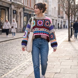 May include: A long-sleeve sweater featuring a multi-colored crochet granny square pattern in shades of pink, blue, yellow, and brown, paired with blue denim jeans, white sneakers, and a plain beige canvas tote bag carried over the shoulder.