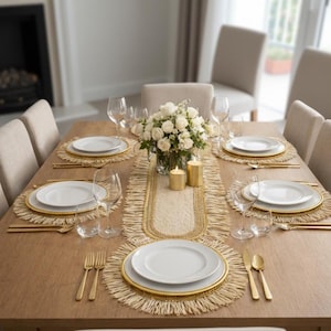 Peut inclure: Une table à manger dressée pour un repas formel. La table est en bois clair et est garnie d'assiettes blanches, de couverts dorés et de sets de table tissés. Une pièce maîtresse florale et des bougies complètent le cadre élégant.