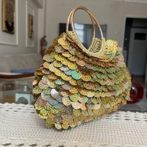 May include: A gold sequined handbag with a circular handle. The bag is covered in iridescent sequins that shimmer with multiple colours. The handle is a matching gold colour. The bag is on a white crocheted table runner.