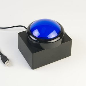 May include: A large, blue button with a black base and a USB cable. The button has a glossy, rounded top and is set on a square, black base. The USB cable extends to the left.