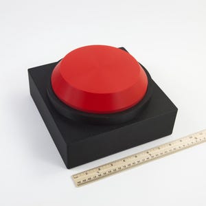 May include: A large, red button on a black square base. A wooden ruler is placed next to the button. The button is approximately 4 inches in diameter. The base is approximately 6 inches square.