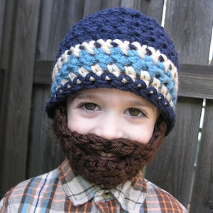 Peut inclure: Un enfant portant un bonnet crocheté bleu, blanc et marron avec une barbe crocheté marron attachée. Le bonnet a une bande rayée blanche et bleue.