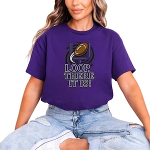 May include: A purple t-shirt with a football-themed graphic. The design features a brown football soaring through a goalpost, with the text "LOOP, THERE IT IS!" in a stylized font. The shirt is made of a soft material.