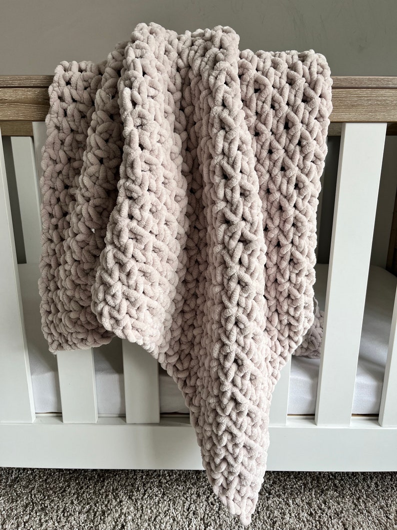 May include: A soft, light pink knitted baby blanket draped over a white crib.