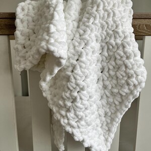 May include: White crocheted baby blanket with a soft, fluffy texture. The blanket is draped over a white crib.