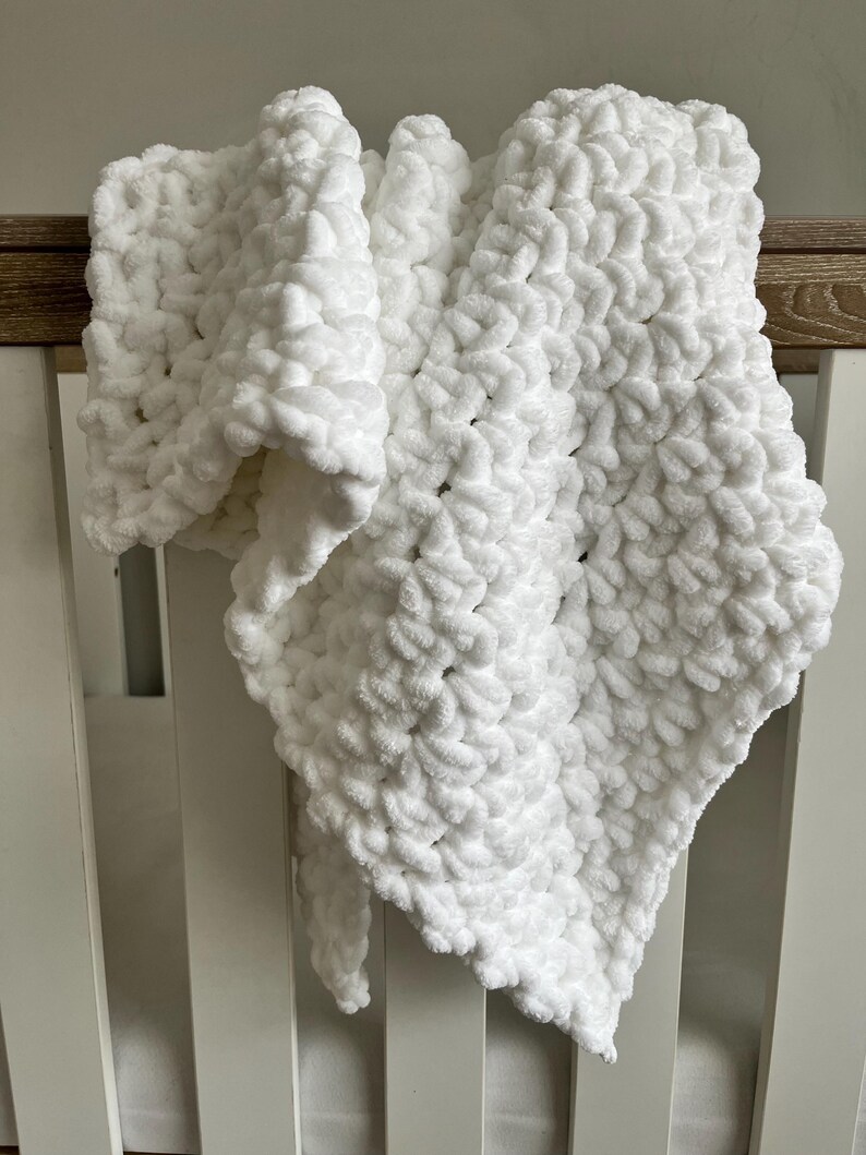 May include: White crocheted baby blanket with a soft, fluffy texture. The blanket is draped over a white crib.