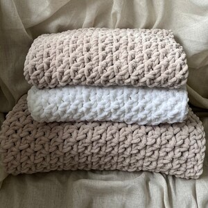 May include: A stack of three knitted blankets in beige and white. The blankets are made of a soft, fluffy yarn and have a textured pattern.