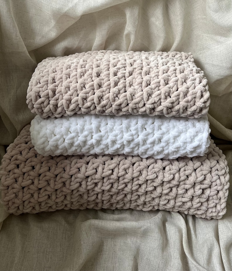 May include: A stack of three knitted blankets in beige and white. The blankets are made of a soft, fluffy yarn and have a textured pattern.