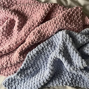 Può includere: Due coperte all'uncinetto, una rosa chiaro e l'altra blu chiaro, sono piegate e sovrapposte. Le coperte sono realizzate con un filato spesso e hanno una texture morbida e soffice.