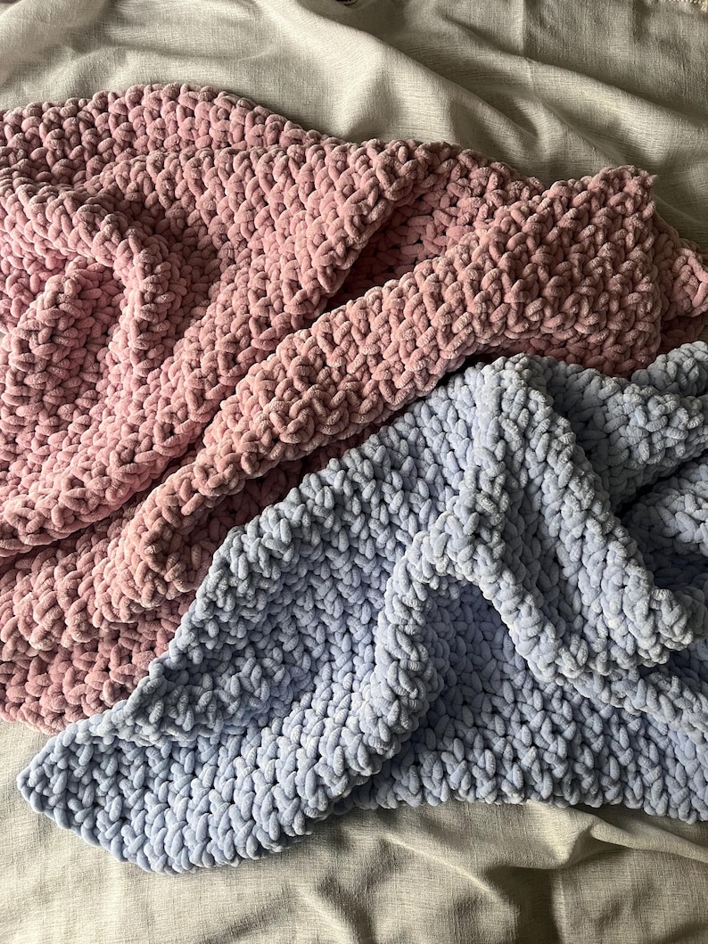 May include: Two crocheted blankets, one in a light pink and one in a light blue, are folded and layered on top of each other. The blankets are made with a chunky yarn and have a soft, fuzzy texture.