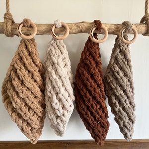 Op de afbeelding: Vier gehaakte babydekens in beige, crème, bruin en grijs, hangend aan een houten tak met houten ringen.
