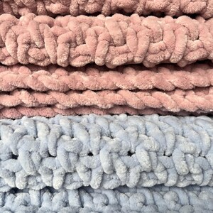 May include: Close-up of a soft, chunky knit blanket in two colors: dusty rose and light blue. The blanket is made with thick yarn and has a textured, woven pattern.