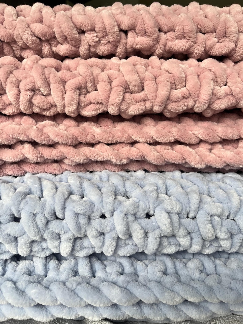 May include: Close-up of a soft, chunky knit blanket in two colors: dusty rose and light blue. The blanket is made with thick yarn and has a textured, woven pattern.