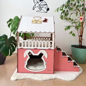Puede incluir: Una casa de mascotas de madera rosa y blanca con un recorte de cara de conejo y escaleras. La casa tiene un techo festoneado y un pequeño letrero que dice "Willow & Leo". Una decoración de pared de conejo geométrico está arriba.