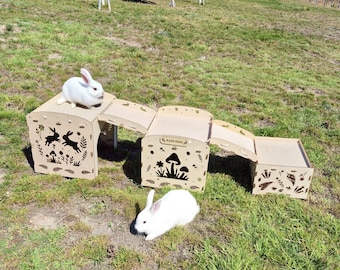 May include: A wooden rabbit play structure with three connected sections. The structure features cut-out designs of rabbits, mushrooms, and foliage. Two white rabbits are present, one on top and one below the structure, on a grassy surface.