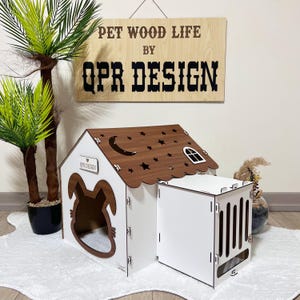May include: White wooden pet house with a brown roof featuring a moon and stars. The house has a cat-shaped entrance and a separate white wooden cage with a slatted door. The sign above reads "PET WOOD LIFE BY QPR DESIGN".
