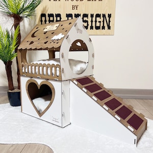 Puede incluir: Una casa para gatos de madera con una entrada en forma de corazón, una rampa y un nivel superior con un diseño de luna y estrellas. La casa está pintada de blanco con detalles marrones. El texto "OPP DESIGN" es visible en la pared detrás de la casa.
