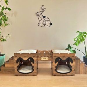 Könnte beinhalten: Ein Haustierbett aus Holz mit Hasenohren-Design. Das Bett hat zwei Fächer mit weißen Kissen. Über dem Bett befindet sich eine geometrische Hasen-Wandkunst. Das Bett steht auf einer Holztreppe.