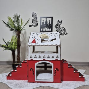 May include: A red and white wooden pet house with a Christmas theme. The house features a sloped roof with snowflake and reindeer designs, a front door, and a staircase. The word "Brownie" is on the roof.