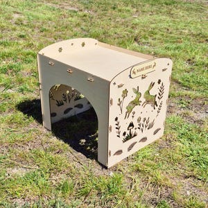 Puede incluir: Un banco de madera de color claro con una parte superior curvada y una abertura arqueada. Los paneles laterales presentan diseños recortados de conejos, flores y follaje. La parte superior tiene pequeños recortes de flores y el texto "NAME HERE".