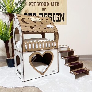 以下が含まれることがあります： 白を基調とし、茶色の屋根が付いた2階建ての木製ペットハウス。前面にはハート型の切り抜きがあり、装飾的な手すりが付いています。階段はバーガンディ色のステップで構成されています。テキスト「PET WOOD LIFE BY OPR DESIGN」が表示されています。