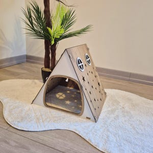 以下が含まれることがあります： 明るい木目の仕上げと白いアクセントが施された、テント型の木製ペットハウス。前面には大きな開口部があり、側面には2つの楕円形の窓があります。テントは星の切り抜きで装飾されており、白くふわふわのラグの上に置かれています。
