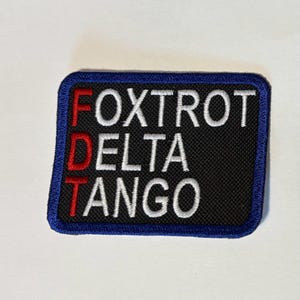 May include: A rectangular patch with the words "FOXTROT DELTA TANGO" embroidered in white, red, and black. The patch has a blue border and is likely designed for application on clothing or gear.