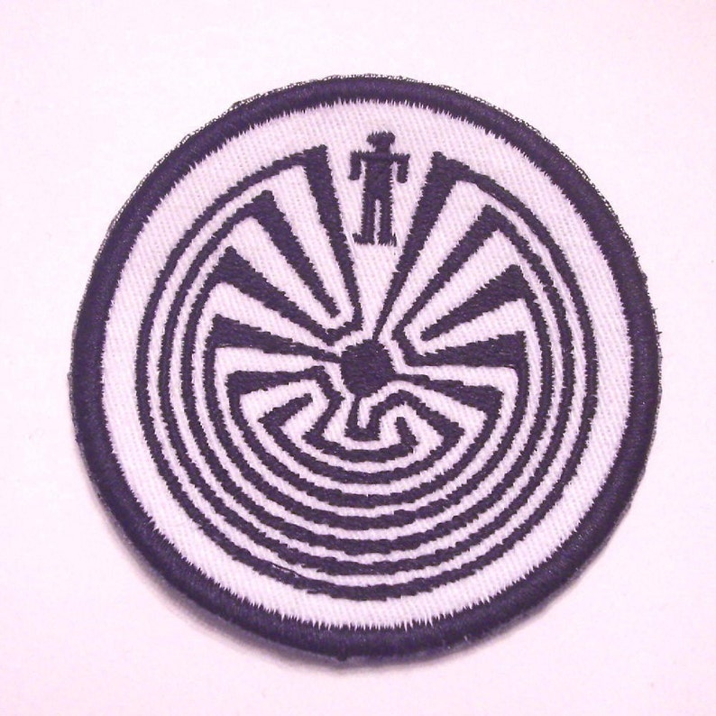 May include: A white circular patch with a black embroidered design of a Native American labyrinth pattern. The center of the labyrinth has a black circle and a small black stick figure.